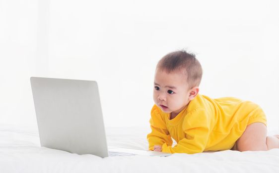 Asian little baby girl wearing a yellow dress typing hands on laptop computer keyboard and looking at monitor on the white bed. Little IT guy