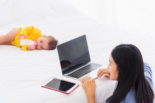 Asian young woman is working busy on laptop computer and tablet at home office in bedroom while her little baby is sleeping feeding lying on the white bed lying near mom