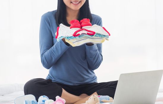 Asian mother shopping online for her baby clothes on laptop computer and tablet on the bed she makes purchase new baby clothes for an unborn baby preparing for the new child