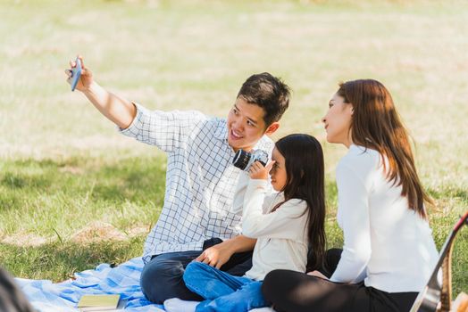 Happy Asian young family father, mother and child little girl having fun and enjoying outdoor sitting on picnic blanket taking selfie using technology mobile smart phone at summer garden park