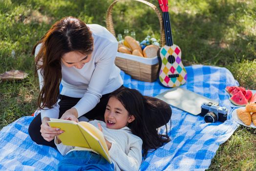 Happy Asian young family mother and child little girl having fun and enjoying outdoor laying on picnic blanket reading book at summer garden spring park, Family relaxation concept