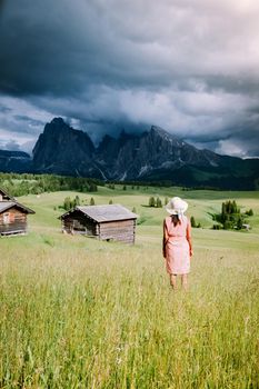 Alpe di Siusi - Seiser Alm with Sassolungo - Langkofel mountain group in background at sunset. Yellow spring flowers and wooden chalets in Dolomites, Trentino Alto Adige, South Tyrol, Italy, Europe. Summer weather with dark clouds rain, couple men and woman on vacation in the Dolomites Italy
