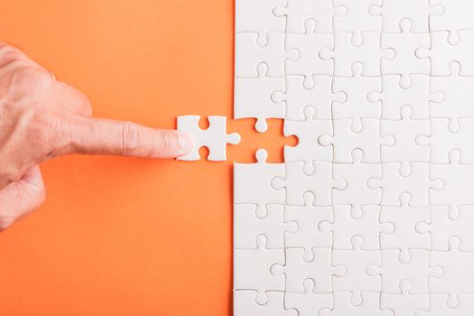 Top view flat lay of hand-holding last piece white paper jigsaw puzzle game last pieces put to place for solve problem complete mission, studio shot on an orange background, quiz calculation concept