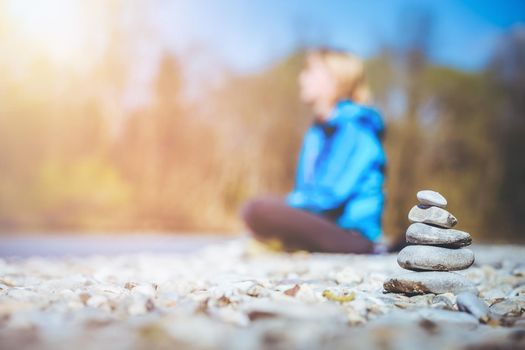 Cairn on a pebble beach, meditating woman in the background. Morning sun.