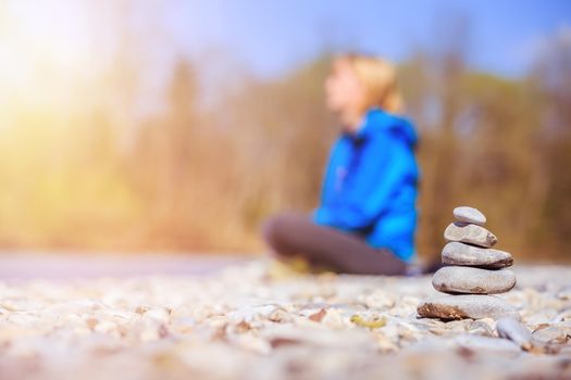 Cairn on a pebble beach, meditating woman in the background. Morning sun.