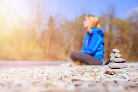 Cairn on a pebble beach, meditating woman in the background. Morning sun.