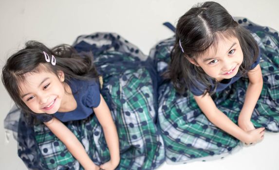 An Identical twin girls sisters are posing for the camera. Happy twin sisters in dresses are looking at the camera and smiling. Frontal view, on white background