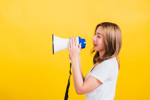 Asian Thai happy portrait beautiful cute young woman stand to make announcement message shouting screaming in megaphone looking to side away, studio shot isolated on yellow background with copy space