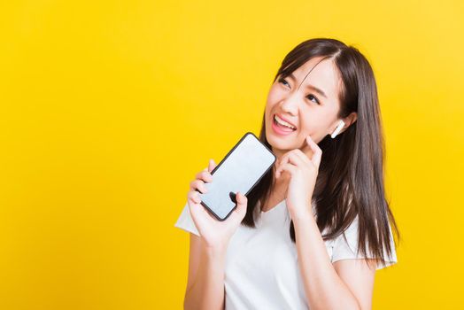 Portrait Asian of a happy beautiful young woman holding a mobile smart phone and wearing wireless headphones listening to music from smartphone studio shot isolated on yellow background