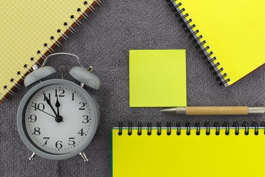 Color trends themed still life with alarm clock placed between of yellow wire bound notepads and pen on a textured grey background viewed as a flat lay from overhead