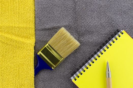 Colorful yellow and grey themed still life with clean paintbrush and yellow wire bound notepad on a two tone grey and yellow textile background