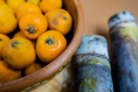 Fresh Mexican hawthorns in handmade clay bowl next to two sugar canes. Close-up of orange hawthorns in brown bowl. Healthy snack preparation