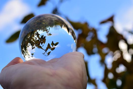 Blue sky with a few clouds and some trees trough a crystal lens ball on a human hand
