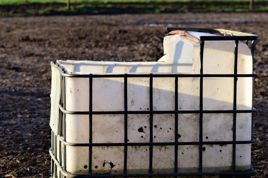 Big white water container with iron grid for stabilisation for animals on a paddock and the leg of a horse in the bachground