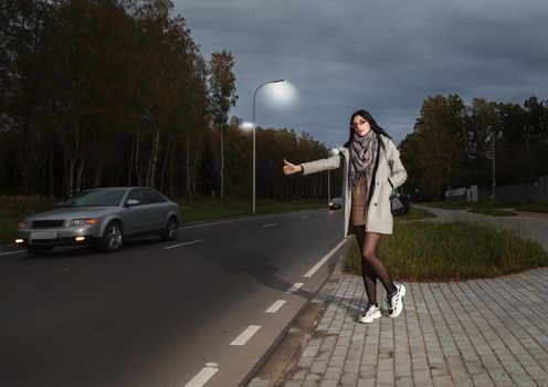 young girl in a gray coat stands near the road trying to stop a car in a late autumn evening