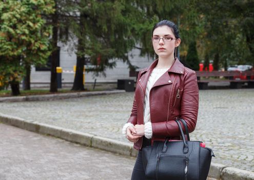 lonely young girl in a brown jacket and black jeans standing on the sidewalk on autumn day