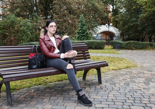 young girl in brown jacket and black jeans sitting on a park bench in autumn