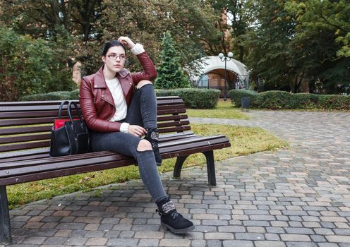young girl in brown jacket and black jeans sitting on a park bench in autumn