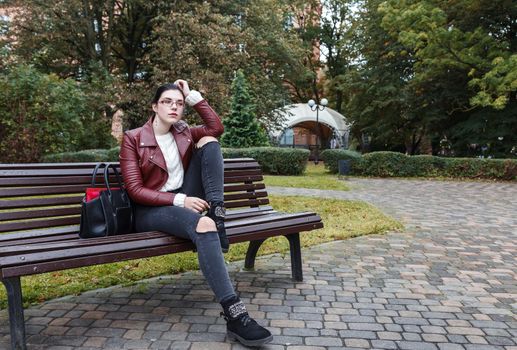 young girl in brown jacket and black jeans sitting on a park bench in autumn