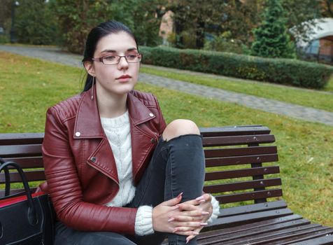 young girl in brown jacket and black jeans sitting on a park bench in autumn. portrait closeup