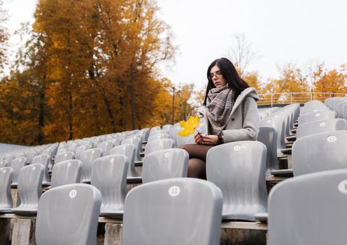 lonely young girl in a gray coat sitting on a seat of an empty stadium on autumn day