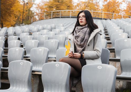 lonely young girl in a gray coat sitting on a seat of an empty stadium on autumn day