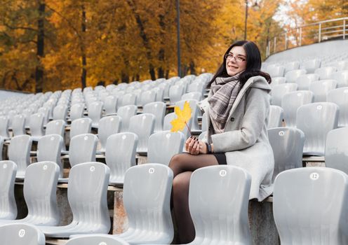 smiling young girl in a gray coat sitting on a seat of an empty stadium on autumn day