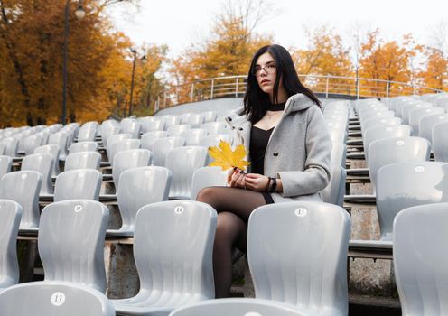lonely young girl in a gray coat sitting on a seat of an empty stadium on autumn day