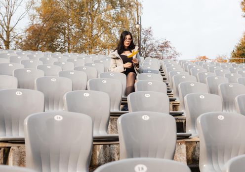 lonely young girl in a gray coat sitting on a seat of an empty stadium on autumn day