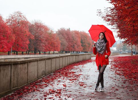 young girl in a red coat with an umbrella stands on the alley of the park after the rain on gloomy autumn day