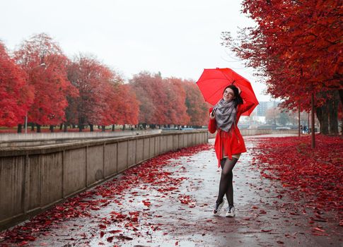 young smiling girl in a red coat with an umbrella stands on the alley of the park after the rain on gloomy autumn day