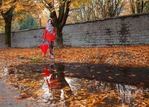 young girl in a red coat with an umbrella stands on the alley of the park after the rain on gloomy autumn day