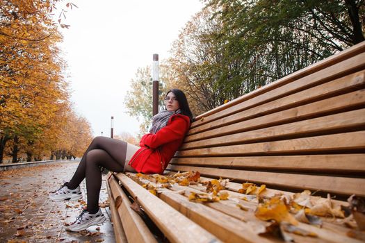 young beautiful girl in a red coat sitting on a bench in a city park after the rain on an autumn day