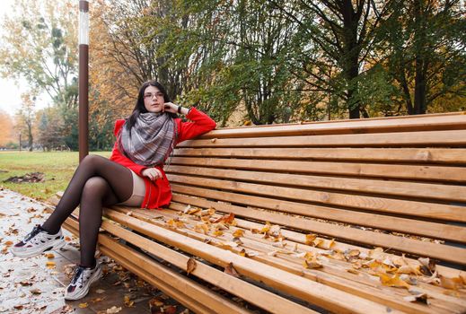 young beautiful girl in a red coat sitting on a bench in a city park after the rain on an autumn day