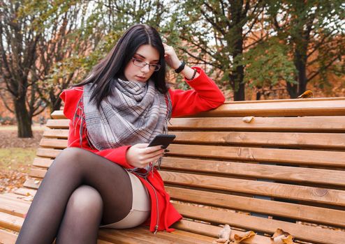 young beautiful girl looking at smartphone display while sitting on a park bench on autumn day