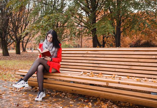 young beautiful girl in red coat reading book while sitting on a park bench on autumn day