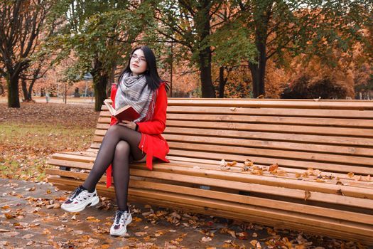 young beautiful girl in red coat reading book while sitting on a park bench on autumn day