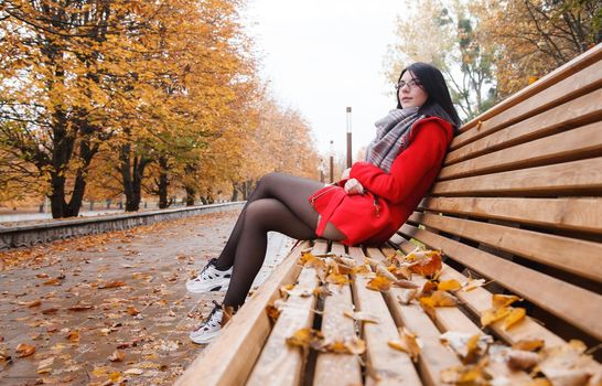 young beautiful girl in a red coat sitting on a bench in a city park after the rain on an autumn day