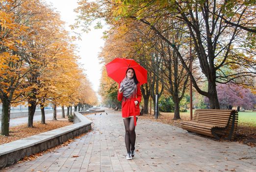 young girl in a red coat with an umbrella walking on the alley of a city park on a sunny autumn day