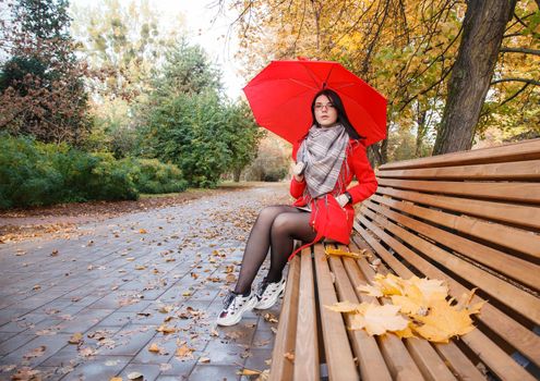 young girl in a red coat with an umbrella sitting on a bench in a city park after the rain on an autumn day