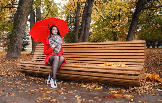 young girl in a red coat with an umbrella sitting on a bench in a city park after the rain on an autumn day