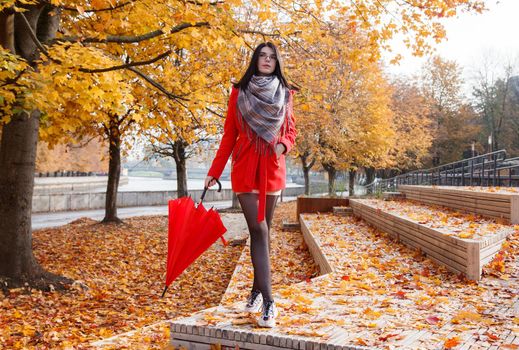 young girl in a red coat with an umbrella standing on the alley of a city park on a sunny autumn day