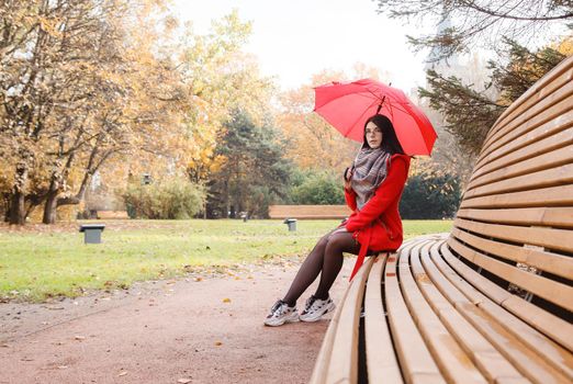 young girl in a red coat with an umbrella sitting on a bench in a city park after the rain on an autumn day