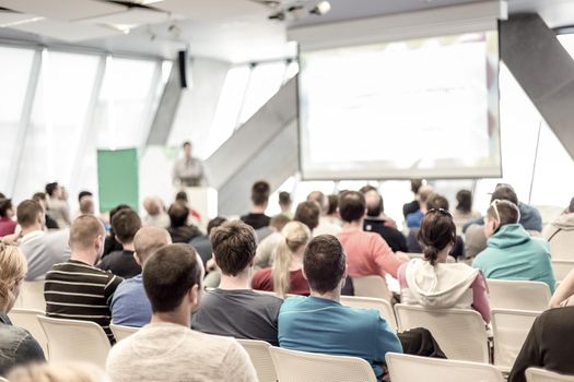 Man giving presentation in lecture hall. Male speeker having talk at public event. Participants listening to lecture. Rear view, focus on people in audience.