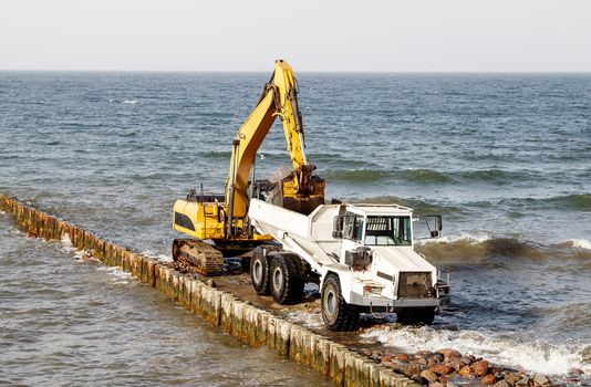 excavator loading soil into a large truck during the construction of a breakwater on the seashore on autumn day