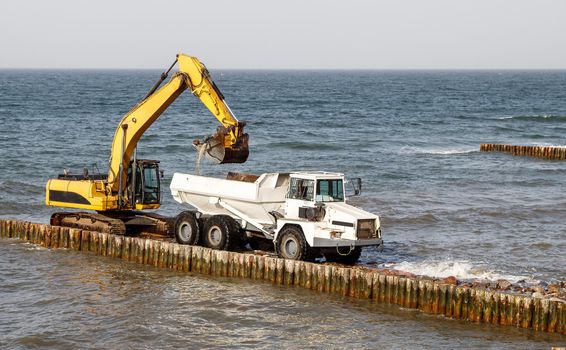 excavator loading soil into a large truck during the construction of a breakwater on the seashore on autumn day
