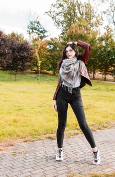 young girl in brown jacket and black jeans standing on the alley in city park on sunny autumn day