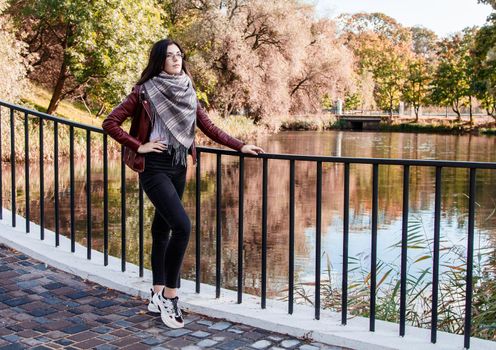 young girl in brown jacket and black jeans standing on bridge near the river in city park on sunny autumn day