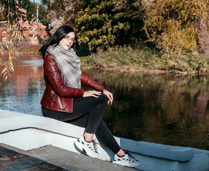 young girl in brown jacket and black jeans sitting on a parapet near a pond in city park on sunny autumn day