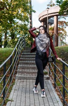 young girl in brown jacket and black jeans standing on the stairs in city park on sunny autumn day
